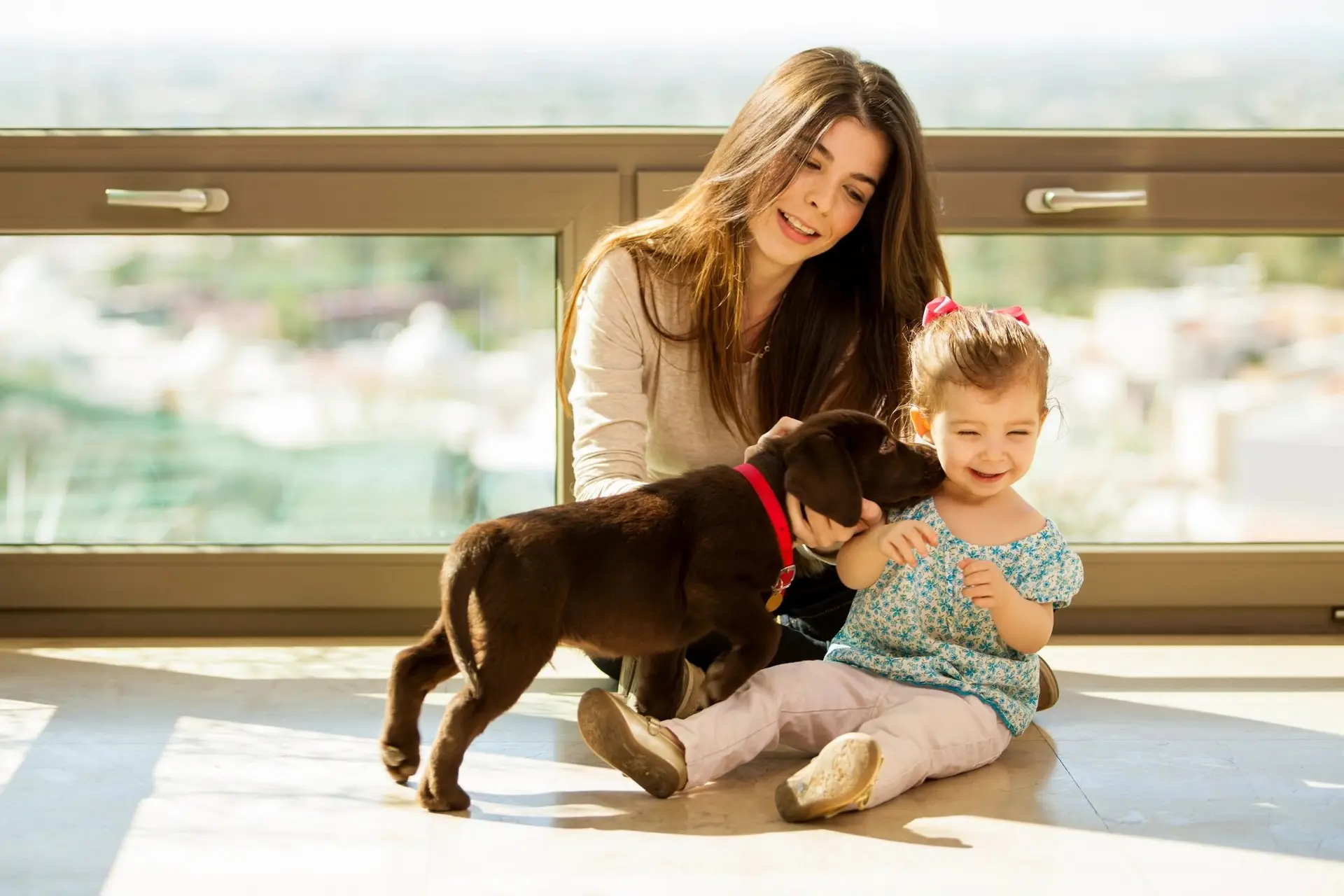 Mother and baby smiling as family dog interacts playfully with the infant.