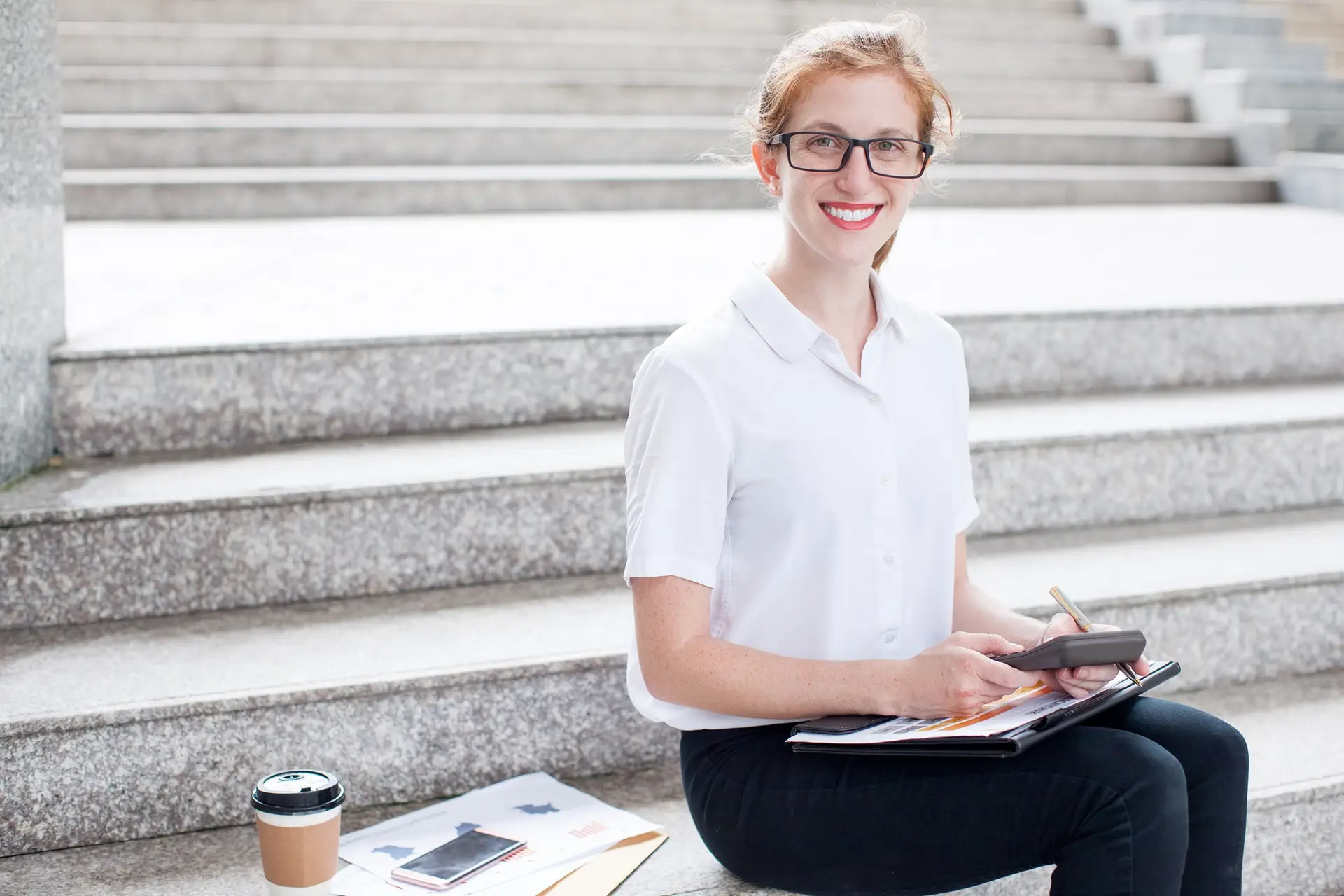 Closeup portrait of smiling attractive young female student looking at camera, using calculator, working and sitting on stairs outdoors